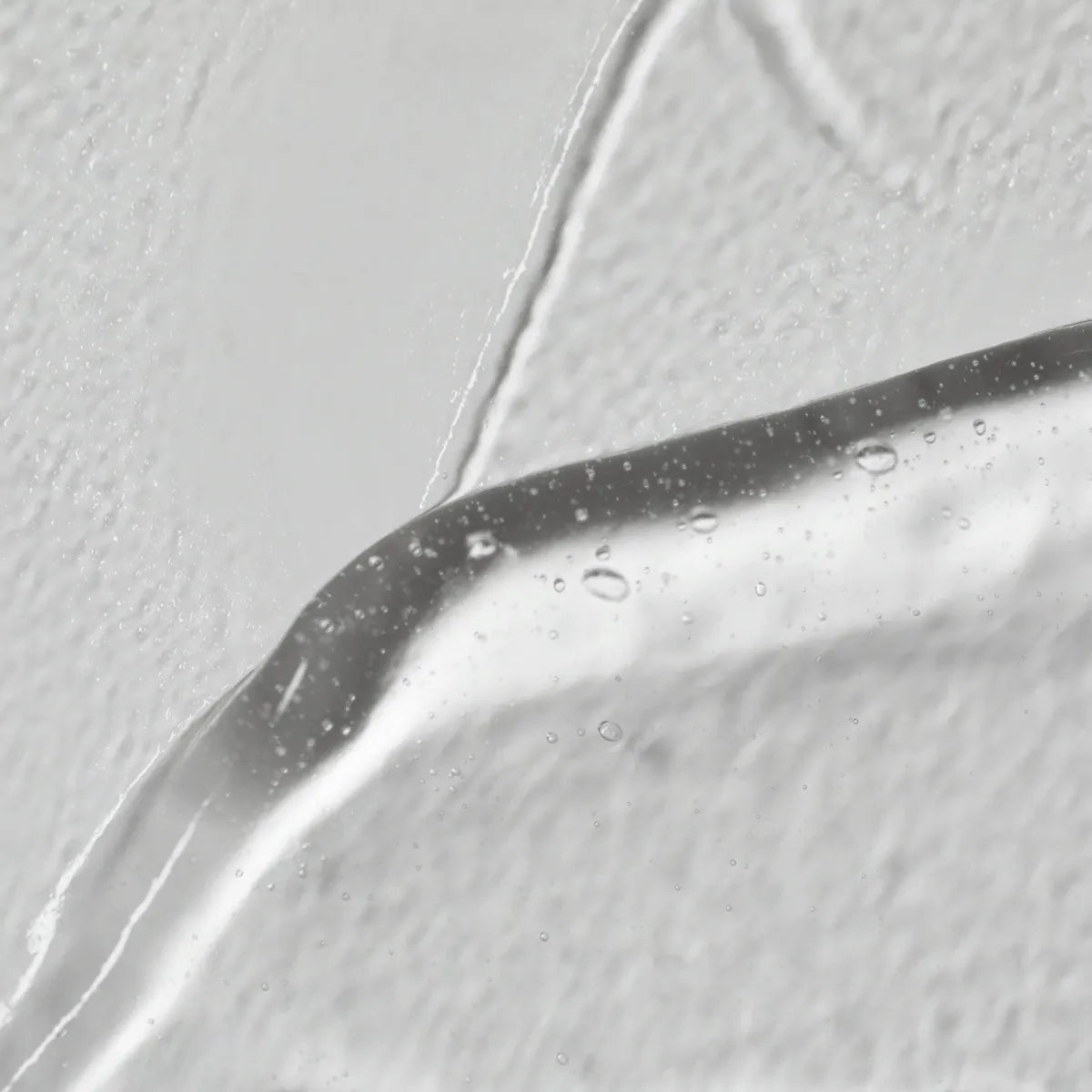 Nahaufnahme von Solawaves LightBoost Wand Activating Serum, einem klaren Gel mit Hyaluronsäure. Es zeigt Luftblasen vor einem strukturierten weißen Hintergrund, der die glatte, glänzende Oberfläche im Kontrast zum rauen Hintergrund hervorhebt.